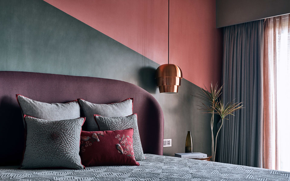 A bedroom with a bed with a large purple headboard, the wall behind painted grey and pink, a hanging brass coloured lamp and a window on the side