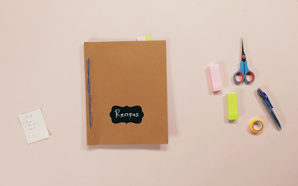 A book with a brown cover with 'recipes' written on it placed on a pink paper with a scissor, a couple of post its, a pen and a tape kept beside the book