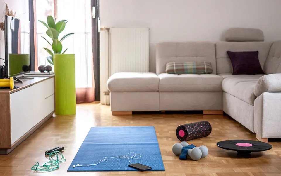 A blue yoga mat, a pair of dumbells, a skipping rope and a phone with headphones lying on the floor of a living room with wooden flooring, a beige coloured L shaped sofa and a Television