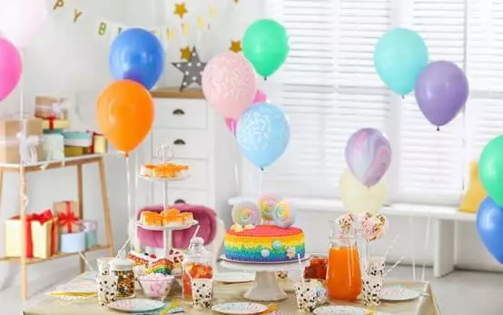 A living room filled with colourful balloons and a table with a cake, jar of orange juice, cups and a cake stand