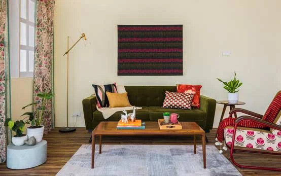 Living room with green sofa, wooden flooring, wooden centre table, large frame ion the wall, side table with a planter, chair with a  red throw on it and a tall golden lamp