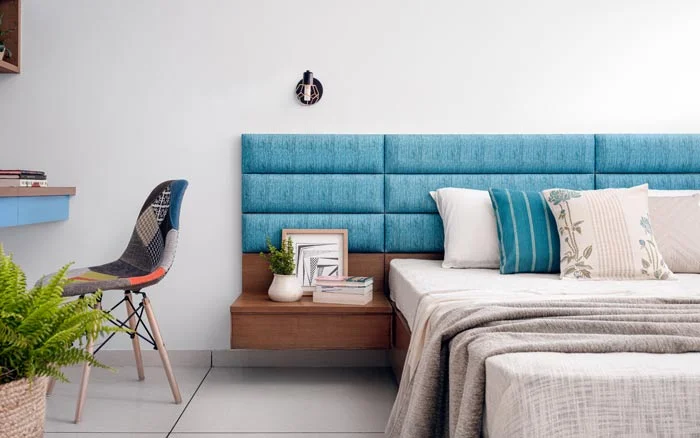 A bedroom with a blue cushioned headboard and a chair placed next to it
