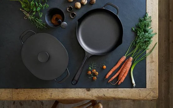 A couple of cast iron utensils kept on a table top along with vegetables like carrots, cherry tomatoes and herbs
