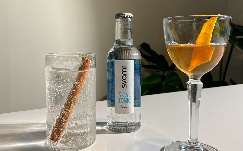 A cocktail glass with yellow liquid and an orange skin, a small bottle with 'Svami' written on it and a glass filled with soda and a stick of cinnamon placed inside it