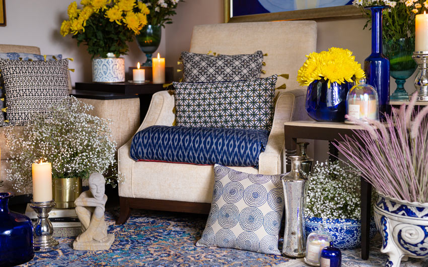 A living room with a beige sofa, along with indigo and grey cushions, Indigo vases, candles with stands and flowers