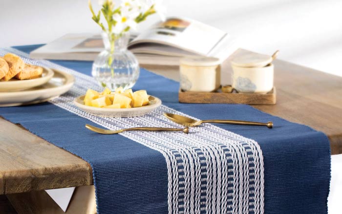 A blue table runner placed on a wooden table with golden spoons, a small vase and ceramic cutlery