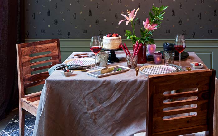 A wooden dining table with a pink table cloth, cutlery placed on it, a flower vase and candles