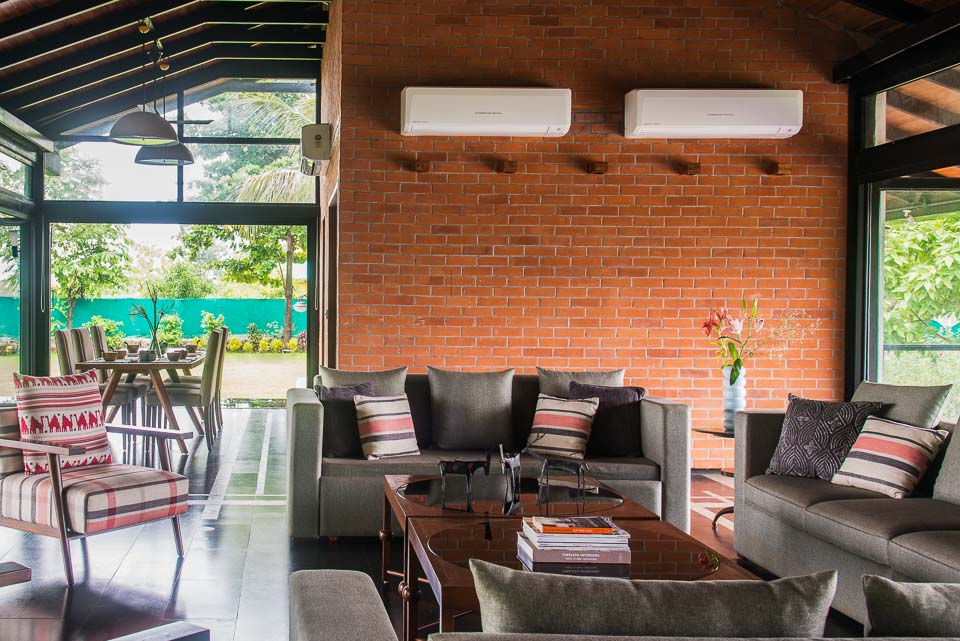 Living room in a farm house in Karjat with two grey sofas, a dining table in the back and with a red bricked wall fitted with two AC&rsquo;s 