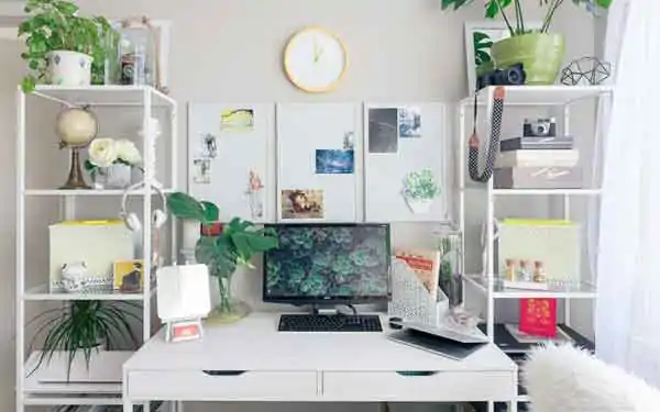 A white Study table in a home with racks to its left and right filled with books, cameras and plants. 